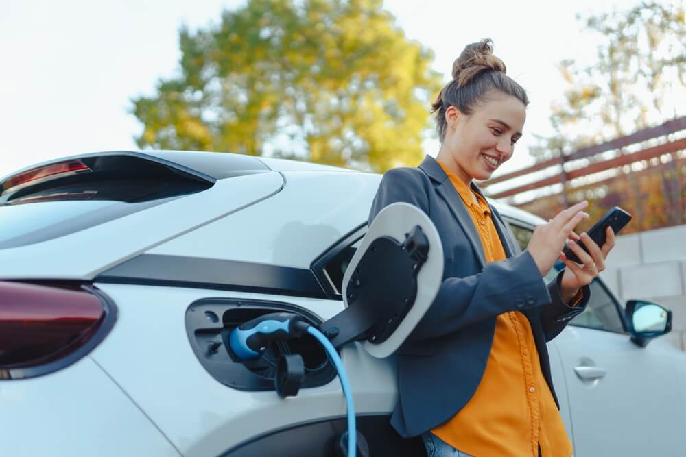 Young woman with smartphone waiting while her electric car charging in home charging station