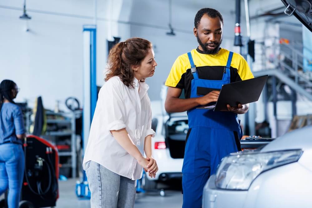 Repairman at auto repair shop conducts yearly vehicle checkup, informing customer about needed engine mending.